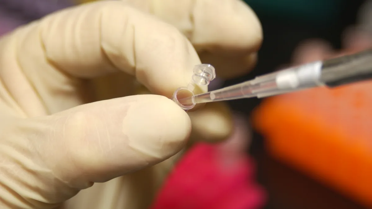 Gloved hands hold a small laboratory tube as a pipette enters its opening.