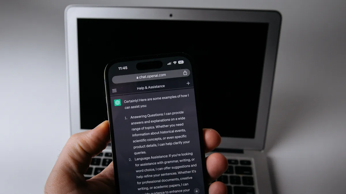 A hand holding a phone displaying ChatGPT in front of an open laptop.