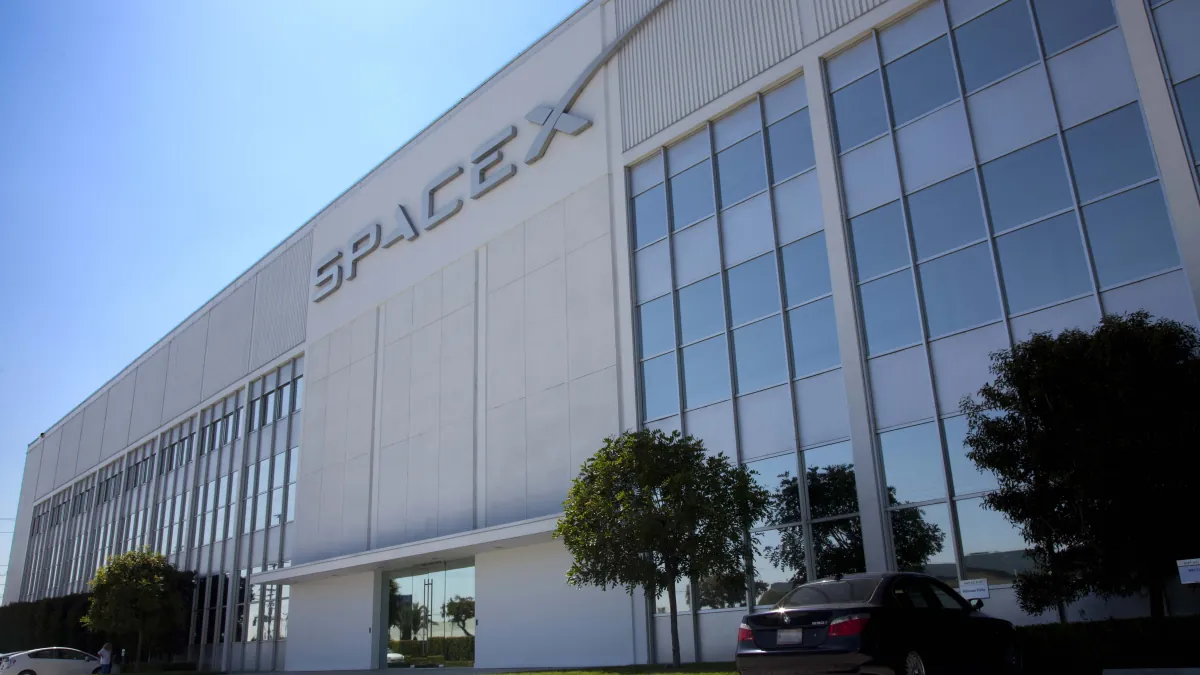 SpaceX lettering on an industrial building with tall windows, trees and parked cars under a blue sky.