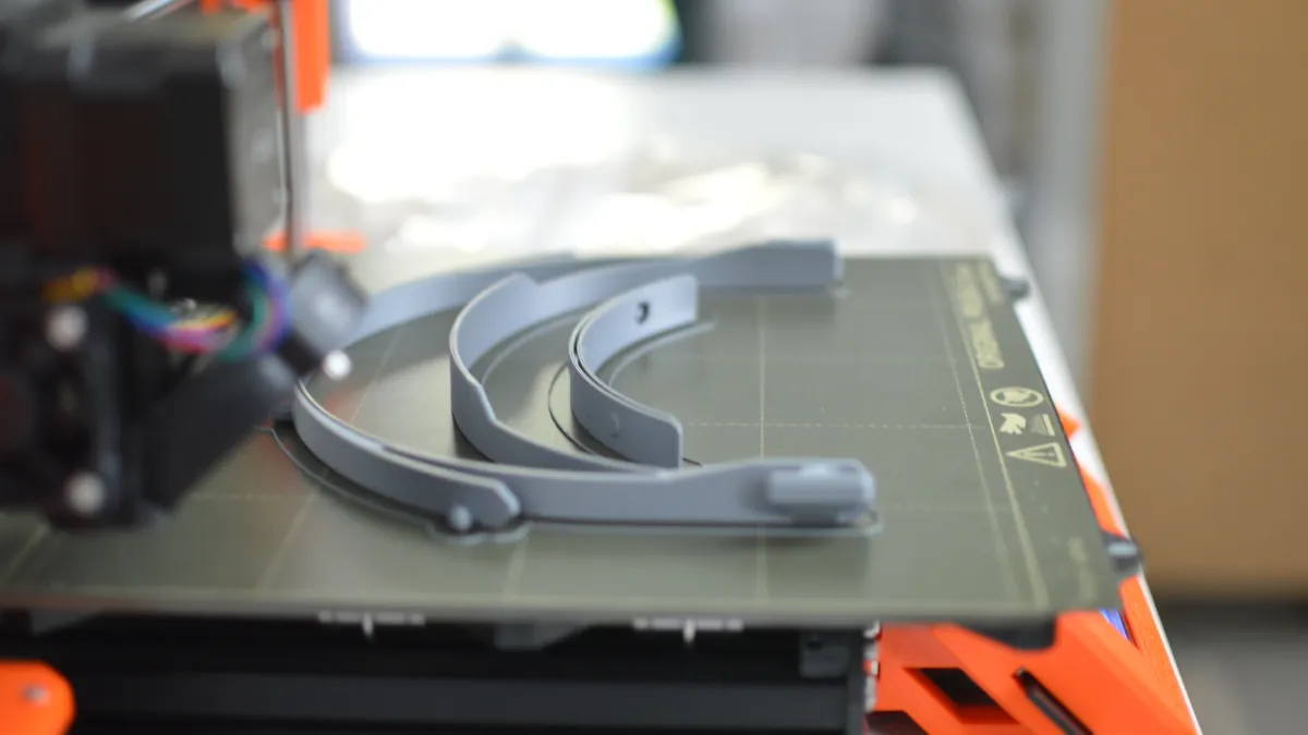 Grey curved faceshield components on a 3D printer bed beneath an orange printer frame.