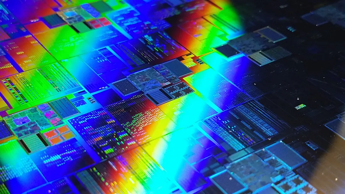 A silicon wafer covered with rectangular chip patterns reflects blue, green and orange light.