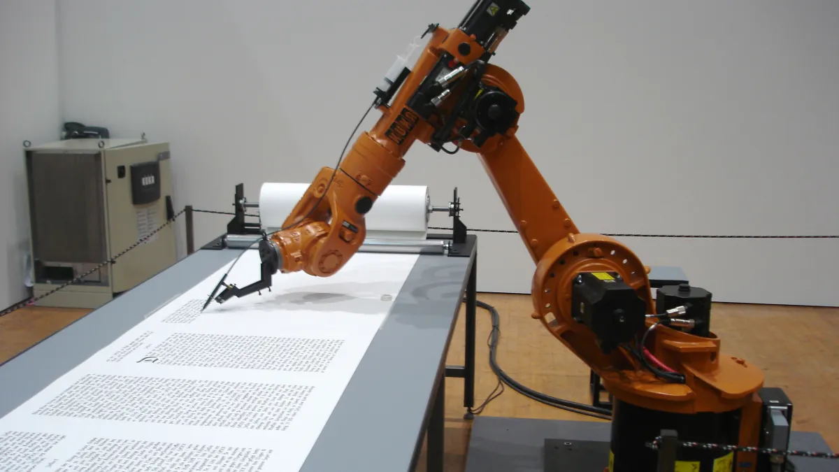 An orange industrial robot holds a pen above a long sheet of handwritten text in an exhibition space.