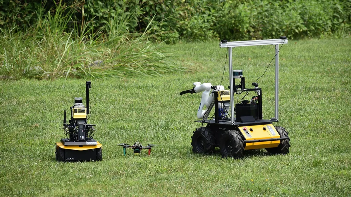 Two yellow-and-black rovers and a small drone on grass; the larger rover carries a robotic arm.