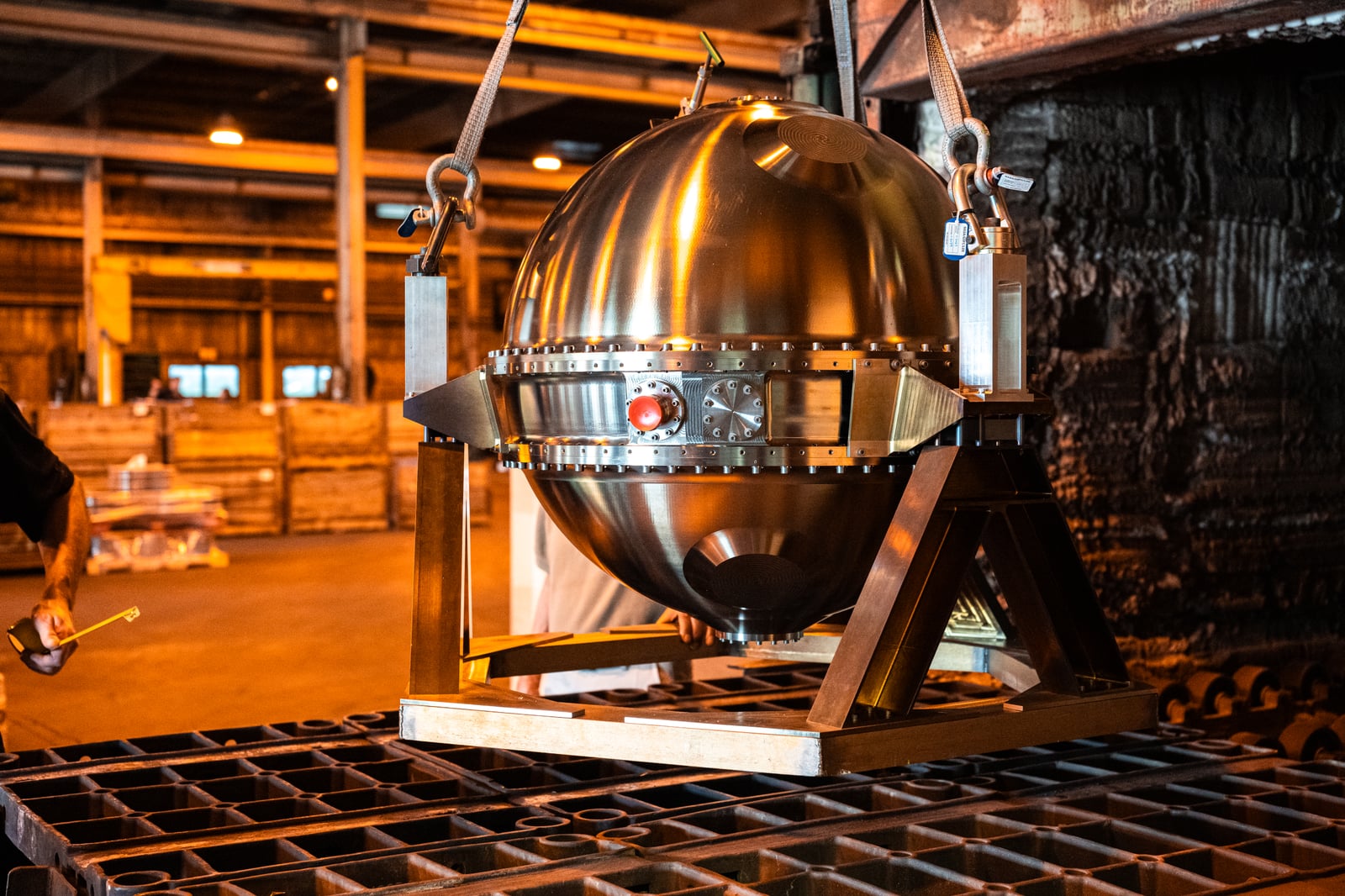 A shiny spherical metal engineering unit suspended by lifting straps above a furnace loading platform.