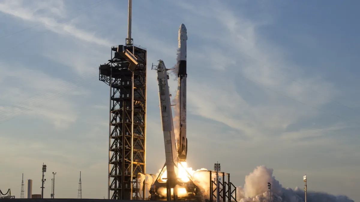 A SpaceX Falcon 9 carrying Crew Dragon lifts off beside its launch tower, with a bright exhaust plume and clouds of smoke.