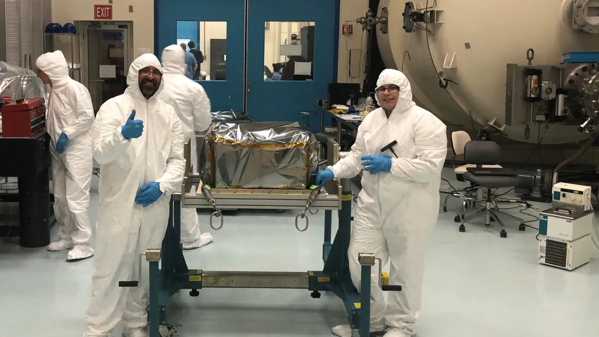 Two researchers wearing cleanroom suits stand beside the foil-covered StarBurst instrument in a NASA testing facility.