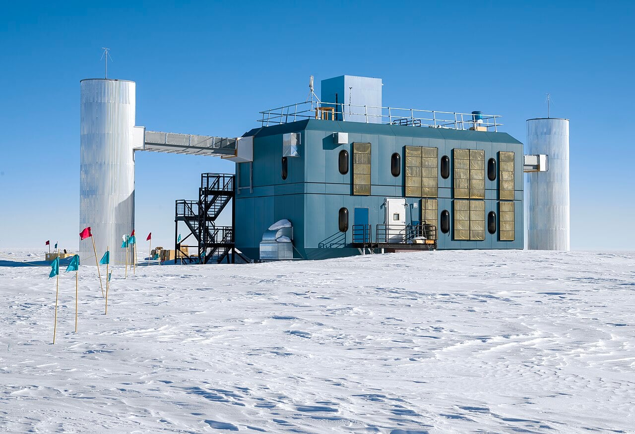 The IceCube surface laboratory stands on Antarctic snow under a clear blue sky, with two silver towers and coloured marker flags.
