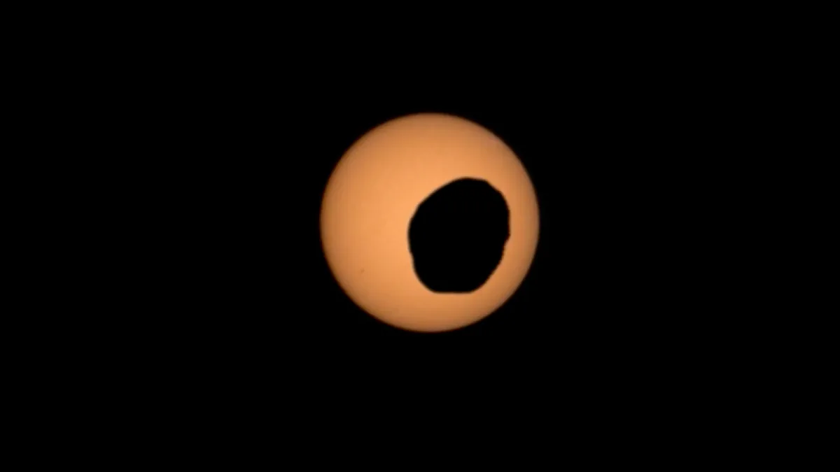 The dark, irregular silhouette of Phobos crossing the orange-tinted solar disc in a Perseverance Mastcam-Z frame.