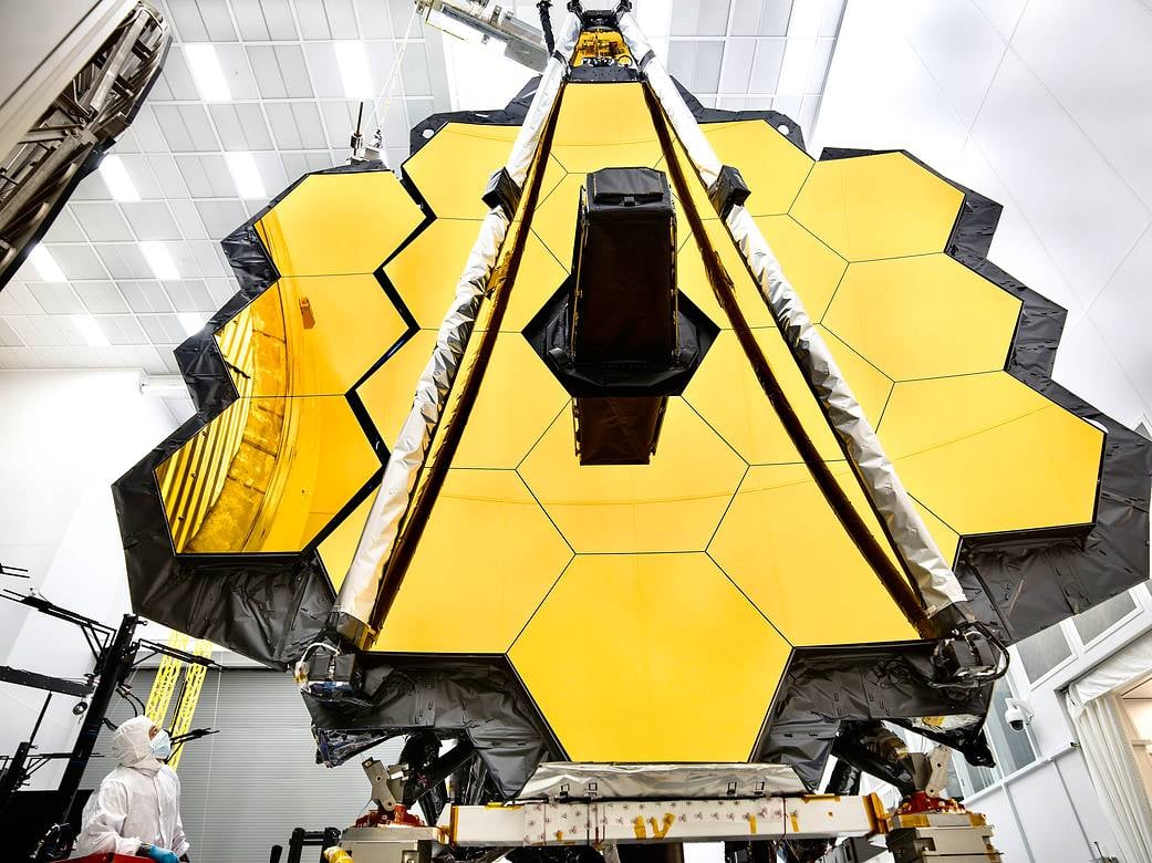 Archive photograph of Webb’s gold segmented primary mirror inside a Johnson Space Center cleanroom, with an engineer watching its deployment.